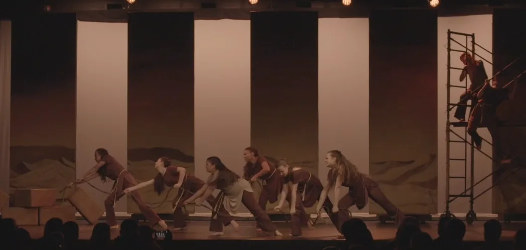Dancers bent low under dim light before tall panels, with scaffolding at stage right.