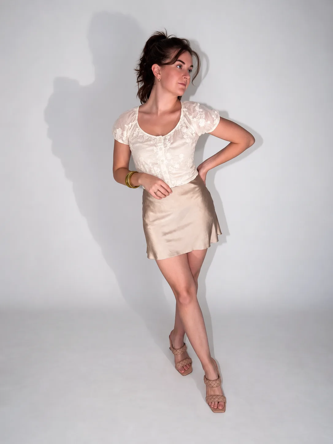 A full-body portrait of a woman in a cream top and champagne skirt with a shadow behind her.